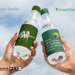 Plakat der Pfandkellnerinnen-Initiative mit zwei erhobenen Mehrwegflaschen vor blauem Himmel. Die Flaschen tragen die Aufschriften „GOLIF“ sowie „Pfandkellnerinnen: Lerchenfelder Straße serviert anders“ und werden von den Logos der Bezirke Josefstadt und Neubau begleitet.