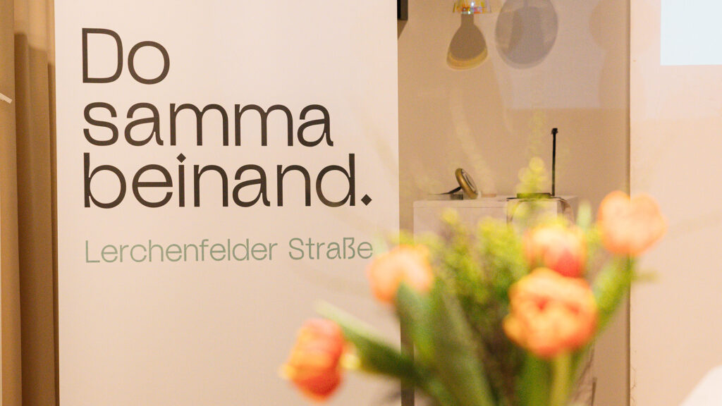 Roll-up der Marke „Do samma beinand“ der IG Kaufleute Lerchenfelder Straße bei der Branding-Präsentation mit unscharfem Blumenarrangement im Vordergrund.